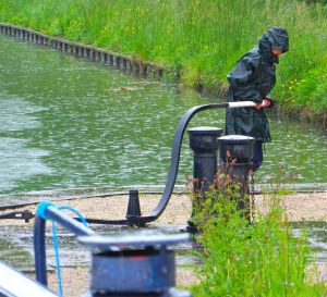 Sandra in full foul weather barging gear opens a manual lock. Harvey is warm and dry in the covered wheelhouse, captaining.